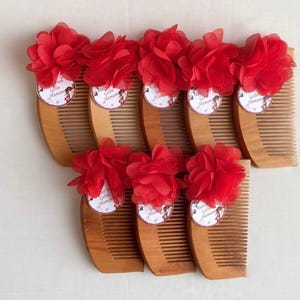 May include: A collection of eight wooden combs, each featuring a large, layered red fabric flower attached to the top. A small, circular white tag with the printed text "Gelin Hamamı" is secured beneath each red flower on the wooden surface.