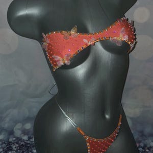 Könnte beinhalten: Ein zweiteiliges Bikini-Set in leuchtendem Orange mit zarten Schmetterlingsapplikationen und verstreuten schillernden Strasssteinen. Das Oberteil ist ein Bandeau-Modell mit dünnen Trägern, das Unterteil ein minimalistischer Tanga mit transparenten Seitenverbindern.
