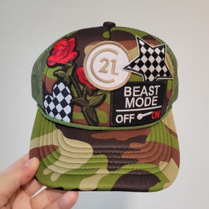 Peut inclure: Casquette de camionneur camouflage avec un dos en filet vert et une visière incurvée. La casquette comporte des écussons brodés, dont une rose rouge, une étoile à damier, un cœur à damier et un écusson avec "BEAST MODE OFF ON".