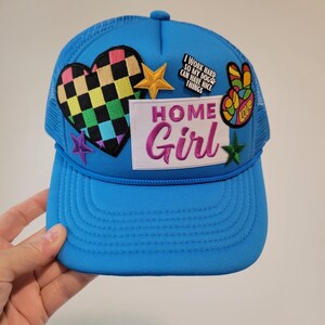 May include: A vibrant blue trucker cap with a mesh back and a curved peak. The front displays patches including a checkered heart, a gold star, a peace sign, and the words "HOME Girl" in pink script. Additional patches include a star and the text "I work hard so my dog can have nice things."