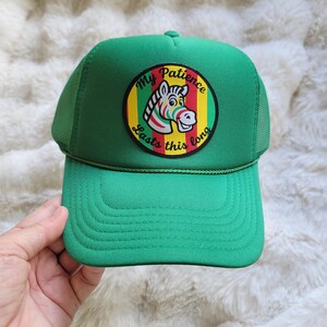 May include: A bright green trucker hat featuring a circular patch on the front. The patch displays a cartoon zebra head with colorful stripes and the text My Patience Lasts this long in black lettering against a red, yellow, and green background.