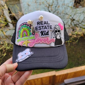May include: A white and black trucker hat with various patches. Patches include a rainbow, the words "REAL ESTATE Kid Lover", and a cartoon image of a woman wearing sunglasses. The hat also has pearl accents.