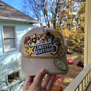 May include: Tan trucker hat with embroidered floral and butterfly patches. The hat features a gold chain detail along the brim and a patch that reads "LIMITED EDITION". A gold star and a "21" patch are also visible.