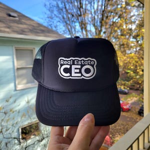 May include: A black trucker hat with a mesh back and a curved brim. The hat features the white text "Real Estate CEO" in a stylized font on the front. The hat is held up against a background of a house and trees.