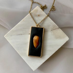 May include: A gold-toned rectangular pendant necklace featuring a smooth black inlay centered with a single, irregular, amber-colored stone. The pendant hangs from a delicate gold-toned chain, creating a contrast between the dark background and the warm, translucent orange-brown gemstone.