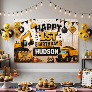 May include: A construction-themed birthday backdrop featuring a yellow excavator and dump truck, with the text HAPPY 1ST BIRTHDAY HUDSON, surrounded by yellow and black balloons, traffic cones, and string lights, creating a festive party scene.