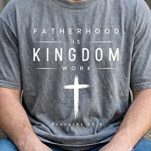 May include: A charcoal gray short-sleeve t-shirt featuring white text that reads FATHERHOOD IS KINGDOM WORK with a white cross graphic below the text and the citation Proverbs 22:6 printed in white at the bottom of the graphic design.