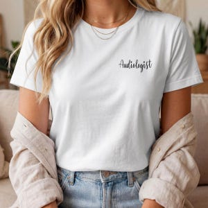 May include: A white short-sleeved t-shirt featuring the word Audiologist printed in black cursive script on the upper left chest area. The shirt is worn with a light-colored button-down shirt layered over it and light blue denim jeans.