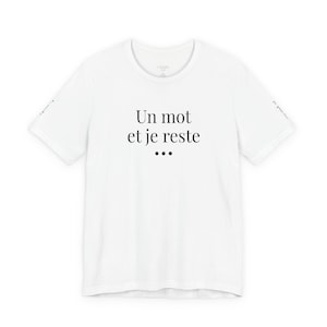 Peut inclure: Un t-shirt blanc à manches courtes avec le texte imprimé en noir 'Un mot et je reste' accompagné de trois points en dessous sur la poitrine. Les manches portent un petit texte noir sur les bords et l'encolure présente une étiquette de marque.