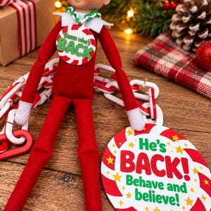May include: A posable elf doll in a red suit with white collar and cuffs. The elf wears a green and white chain necklace with a round tag reading Guess who's BACK. A larger round sign reads He's BACK! Behave and believe! with red and white stripes.