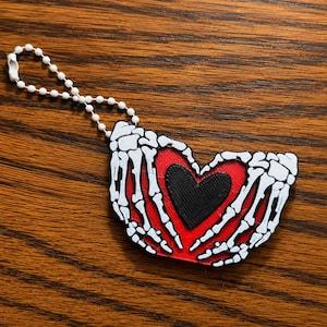 May include: A keychain featuring a 3D-printed design of two white skeleton hands forming a heart shape around a solid black heart, all set against a textured red background. The ornament is attached to a white plastic ball chain connector.
