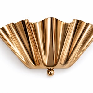 May include: A polished gold-toned wall light featuring a wide, fan-shaped shade with deep, vertical fluted pleats. The fixture is anchored at the base by a single, smooth, spherical gold-toned knob, creating a symmetrical, semi-circular silhouette.