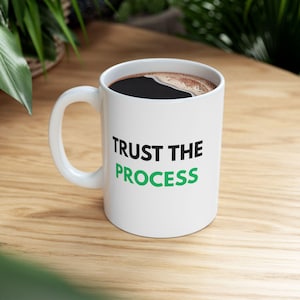 May include: White ceramic coffee mug with the words "TRUST THE PROCESS" printed in black and green. The mug is filled with dark coffee and sits on a wooden surface.