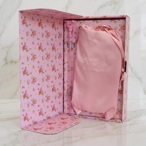 Op de afbeelding: Een roze rechthoekige geschenkdoos met bloemenpatroon met een lichtroze satijnen trekkoordzakje en een doorzichtig rietje met een roze strik. Het zakje is voorzien van de geborduurde tekst STANLEY LOVESHACKFANCY op de voorzijde.
