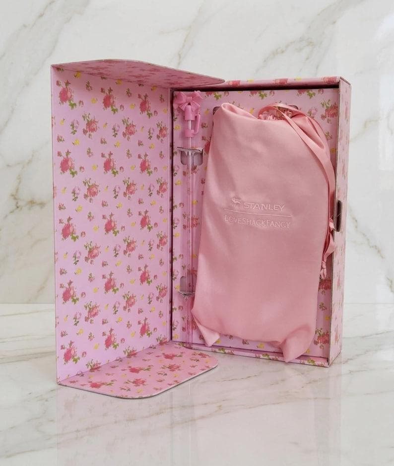 Op de afbeelding: Een roze rechthoekige geschenkdoos met bloemenpatroon met een lichtroze satijnen trekkoordzakje en een doorzichtig rietje met een roze strik. Het zakje is voorzien van de geborduurde tekst STANLEY LOVESHACKFANCY op de voorzijde.