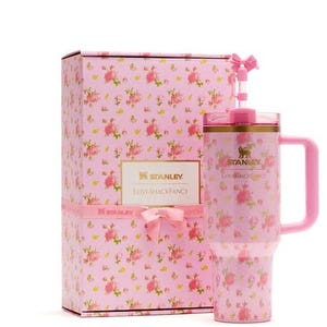 Op de afbeelding: Een roze Stanley LoveShackFancy-beker met een bloemenpatroon, een bijpassend handvat en een rietje met een kleine roze strik. De beker staat naast een rechthoekige geschenkdoos met hetzelfde roze bloemendesign en de merknaam erop.