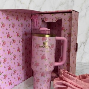 Op de afbeelding: Een roze roestvrijstalen beker met een bloemenpatroon, voorzien van een gouden Stanley-logo en de tekst LoveShackFancy. De beker heeft een handvat, een doorzichtig deksel met een roze bovenkant en zit in een bijpassende doos met een roze stoffen accessoire.