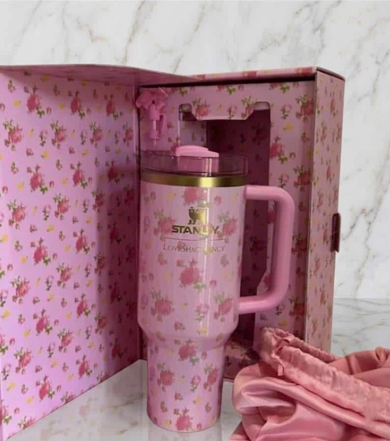 Op de afbeelding: Een roze roestvrijstalen beker met een bloemenpatroon, voorzien van een gouden Stanley-logo en de tekst LoveShackFancy. De beker heeft een handvat, een doorzichtig deksel met een roze bovenkant en zit in een bijpassende doos met een roze stoffen accessoire.