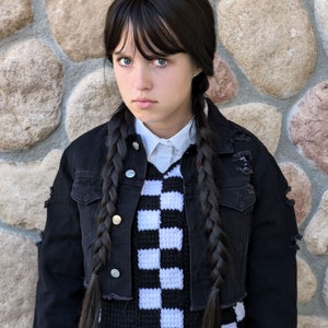 Peut inclure: Une jeune personne portant un gilet en tricot à carreaux noir et blanc sur une chemise blanche boutonnée. Elle porte également une veste en jean noire avec des détails usés et un pantalon noir.