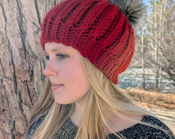May include: A red knit hat with a gray faux fur pom-pom. The hat is ribbed and appears to be handmade. The person in the image is wearing a gray sweater. The background includes a tree trunk and a winter landscape.