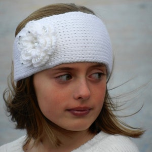 Puede incluir: Una diadema de crochet blanca con un adorno de flor blanca. La flor tiene pequeños diamantes de imitación transparentes.