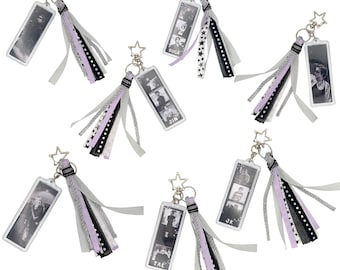 May include: A collection of seven rectangular acrylic keychains, each featuring a star-shaped clasp and a multi-textured tassel. The tassels consist of ribbons in shades of black, white, silver, and lavender. Each keychain displays a black and white photo strip.