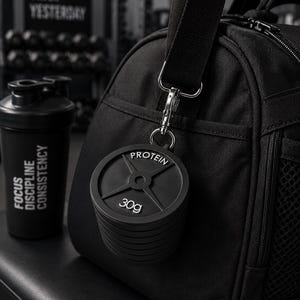 May include: A black gym bag with a silver metal clip attached to a black weight plate keychain featuring the text PROTEIN and 30g. Next to the bag is a black shaker bottle with the text FOCUS DISCIPLINE CONSISTENCY printed in white letters.