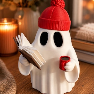May include: A white ghost figurine wearing a bright red knit-style beanie with a pom-pom. The ghost has two black oval eyes, holds an open book with white pages in one hand, and a small red mug filled with a dark liquid in the other hand.