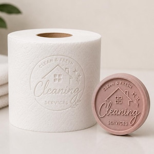 Könnte beinhalten: Eine weiße Toilettenpapierrolle mit einem eingeprägten kreisförmigen Logo, das ein Haussymbol, den Text "CLEAN & FRESH" und "Cleaning SERVICES" zeigt, neben einer passenden runden, malvenfarbenen Wachs- oder Seifenscheibe.