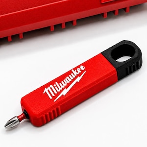 May include: A red and black Milwaukee screwdriver bit holder with a rectangular handle, a circular hole at the top, and a metallic Phillips head bit protruding from the bottom. The brand name Milwaukee is printed in white on the red handle.