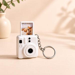 May include: A white miniature camera keyring featuring a square screen on top displaying a photograph of a tan dog. The camera body has a circular lens, two small circular buttons, and a silver metal chain with a circular ring attached to the side.