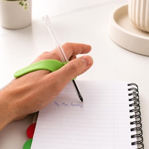 Może przedstawiać: Limonkowy, ergonomiczny uchwyt na długopis przymocowany do przezroczystego długopisu. Długopis jest ustawiony tak, aby pisać w białym notatniku spiralnym w linie. Tekst My Mini Factory jest napisany niebieskim tuszem na stronie notatnika.
