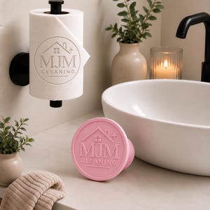 May include: A white paper towel roll mounted on a black holder, featuring a circular logo with a house icon and the text MJM CLEANING. Beside it sits a round, light pink stamp or press tool displaying the same MJM CLEANING logo and house icon.