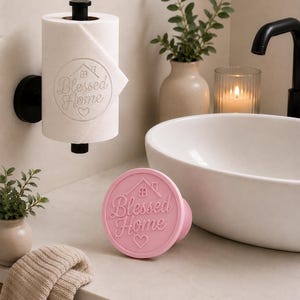 Può includere: Un rotolo di carta igienica bianco montato su un supporto a parete nero, caratterizzato da un design circolare stampato con un'icona di casa, un cuore e il testo Blessed Home. Un timbro circolare rosa abbinato con lo stesso design è sul piano.