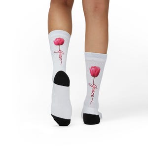 Puede incluir: Calcetines blancos con talón y puntera negros, con un tulipán rosa y la palabra "Grace" en cursiva. Los calcetines son hasta la rodilla y tienen un puño acanalado.
