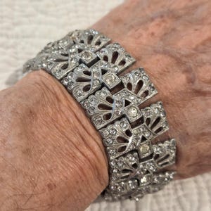 May include: A silver-toned metal link bracelet featuring a repeating fan-shaped design. Each articulated segment is embellished with numerous small, clear, round-cut rhinestones and a single larger, central clear rhinestone in a square setting.