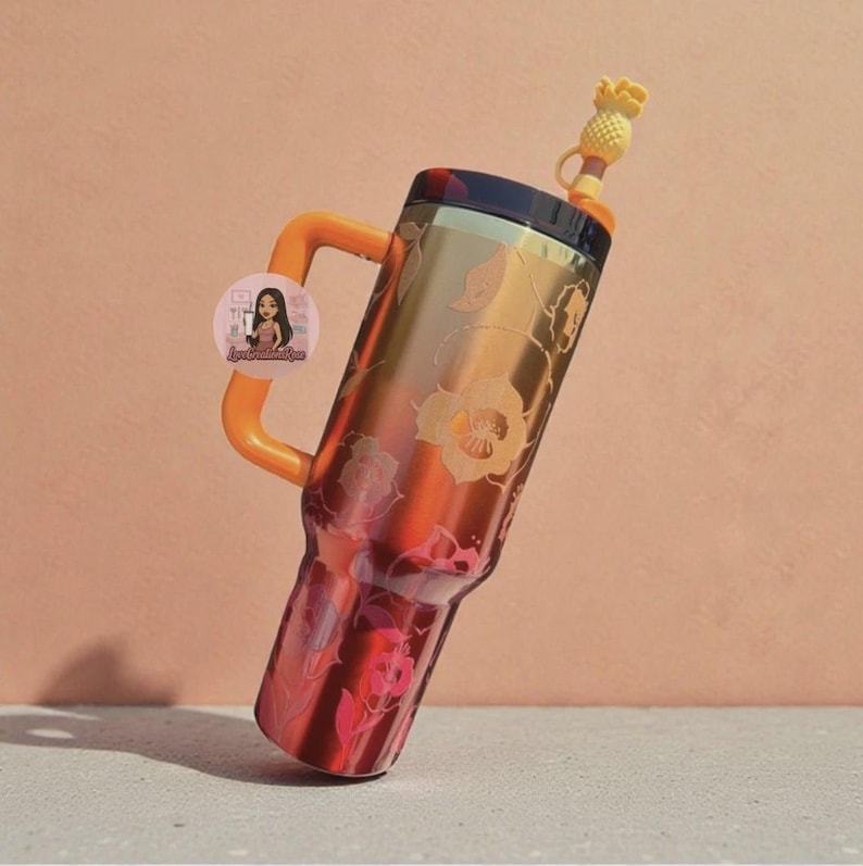 Op de afbeelding: Een hoge, metallic oranje en goudkleurige beker met een handvat, voorzien van een bloemenpatroon en een donker deksel. Een gele ananasvormige rietjesversiering zit op het rietje. Een ronde sticker met een illustratie bevindt zich op het handvat.