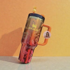 Op de afbeelding: Een metallic oranje Stanley-beker met een donkerbruin deksel, een bijpassend oranje handvat en een gele rietjes-topper in de vorm van een ananas. De buitenkant is versierd met een herhalend bloemenpatroon en het Stanley-logo in gele tekst.