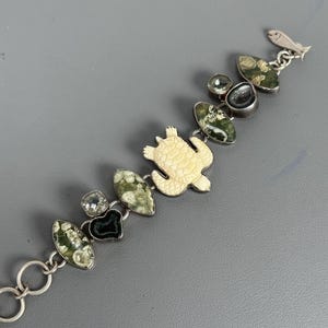 Könnte beinhalten: Ein silberfarbenes Gliederarmband mit einem zentralen cremefarbenen Schildkrötenanhänger, abwechselnd mit ovalen grün gemusterten Steinen, kleinen klaren facettierten Kristallen und dunklen schwarzen Druzy-Steinen, abgeschlossen mit einem Fischverschluss.