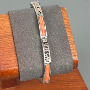 Könnte beinhalten: Ein silberfarbenes Gliederarmband mit abwechselnden rechteckigen Segmenten. Das Design besteht aus glatten, korallenfarbenen rechteckigen Einlagen und durchbrochenen Metallabschnitten mit einem texturierten, unregelmäßigen Muster.