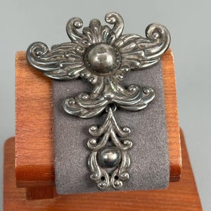 May include: A silver-toned metal brooch featuring an ornate, symmetrical design with scrolling filigree patterns and two smooth, spherical accents. The upper section is wide with wing-like curves, while a smaller, dangling pendant piece hangs below.
