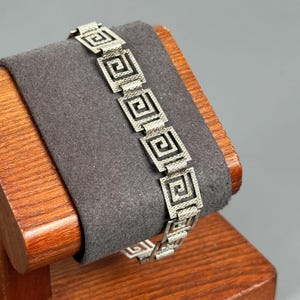 Puede incluir: Una pulsera de metal plateado con un patrón repetitivo de grecas cuadradas. Los eslabones están conectados en una secuencia lineal, cada uno mostrando una superficie texturizada con un diseño de espiral geométrica centrado en un marco cuadrado.