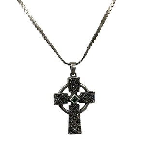 May include: A silver-toned Celtic cross pendant necklace featuring a textured, dark metallic finish with a small, square-cut green gemstone set in the center. The cross is suspended from a matching silver-toned chain with a flat, interlocking link design.
