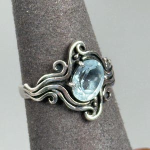 May include: A silver ring featuring an oval-cut, light blue gemstone set in a prong setting. The band is crafted with intricate, swirling metalwork that extends from the central stone, creating a decorative, organic pattern around the finger.