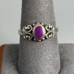 May include: A silver ring featuring a central oval-shaped purple stone set in a bezel. The band is decorated with intricate scrollwork and filigree patterns on both sides of the stone, creating a detailed metal design around the purple center.
