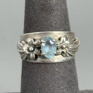 May include: A wide silver band ring featuring a central oval-cut light blue gemstone held by four prongs. The metal band is decorated with sculpted floral motifs on both sides of the stone, showcasing a polished finish with visible metallic texture.