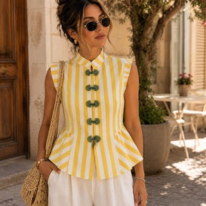 Può includere: Un top peplum senza maniche con righe verticali gialle e bianche, collo alla coreana e cinque chiusure decorative a alamaro verdi sul davanti. Il top è abbinato a pantaloni bianchi e a una borsa a tracolla in paglia intrecciata.