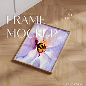 Op de afbeelding: Een rechthoekige houten fotolijst met een aquarelprint van een paarse en witte bloem met een fel oranje en zwart hart. De lijst staat in een hoek, waardoor het kunstwerk duidelijk zichtbaar is binnen de lichte houten rand.