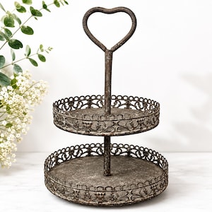 May include: A two-tiered, rustic metal serving tray with a heart-shaped handle. The trays have ornate, scrolled edges and a distressed finish. The handle is made of twisted metal. Perfect for displaying desserts or small items.
