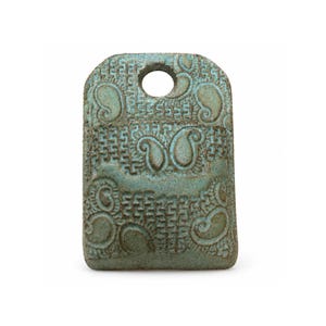 May include: A rectangular ceramic pendant with a rounded top edge and a circular hole for suspension. The surface features a textured, matte teal and brown glaze with an embossed pattern of repeating paisley shapes and geometric grid lines.